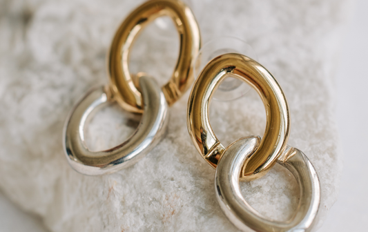Close-up of mixed metal statement earrings featuring interlocking gold and silver links on a textured white stone background.